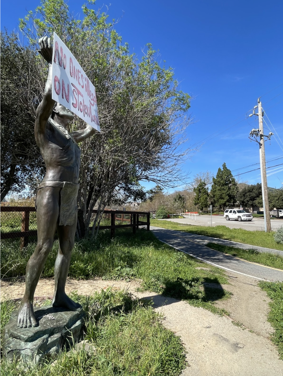 A sign reading “no one’s illegal on stolen land” was placed in the arms of the bronze statue of an indigenous person on the corner of South Higuera Street and Prado Road on Feb. 25.